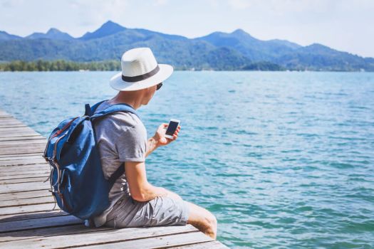 Image shows a traveler sitting on a dock holding a phone. The weather is nice and the sea calm.