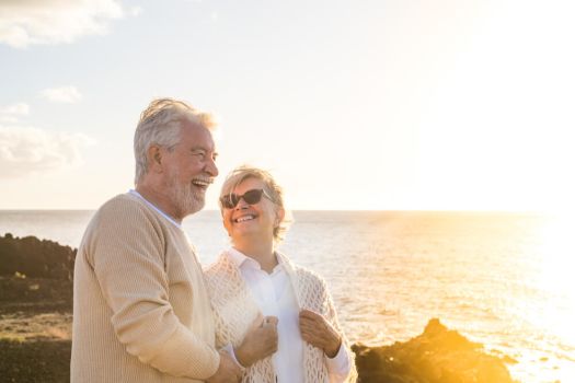 Picture shows a smilling older couple, sharing a joke on a sunny beach as the sun sets. The woman is looking up adoringly at the man.