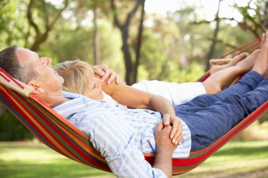 Picture shows an older couple cuddling on a hammock in the sun. They are relaxed and stress free.