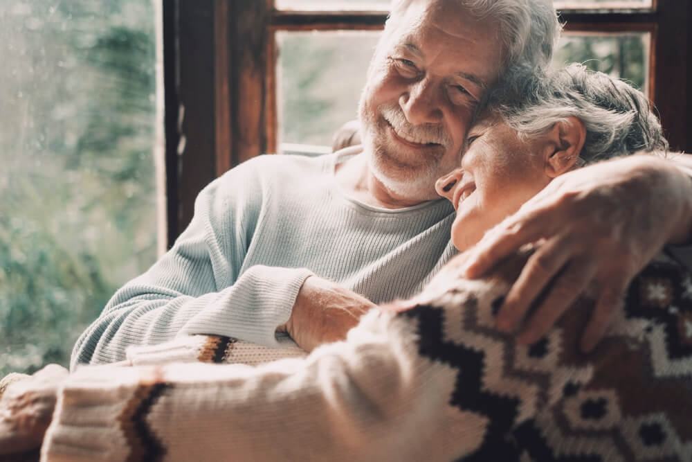 Picture shows a mature couple cuddling and wearing cosy jumpers. Both are smiling.